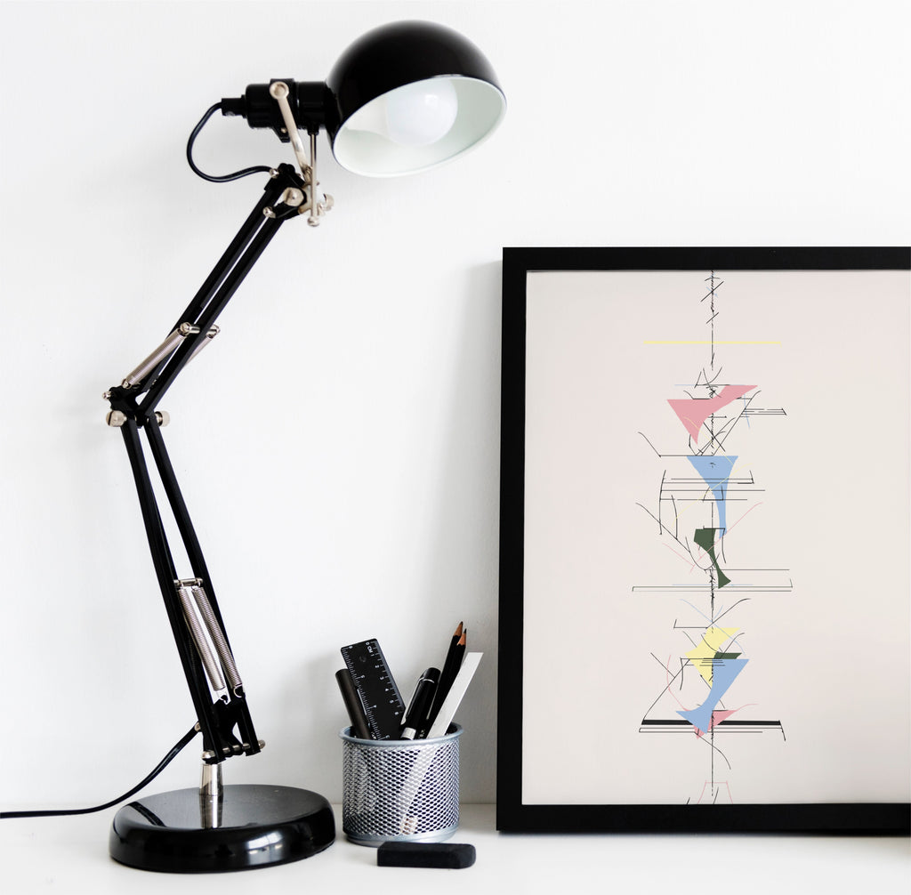 A black adjustable desk lamp with a white interior shade sits on a white surface next to a framed abstract art print. The lamp has a black base and silver metal joints, with a pen cup holding pens nearby.