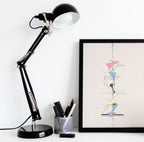 A black adjustable desk lamp with a white interior shade sits on a white surface next to a framed abstract art print. The lamp has a black base and silver metal joints, with a pen cup holding pens nearby.