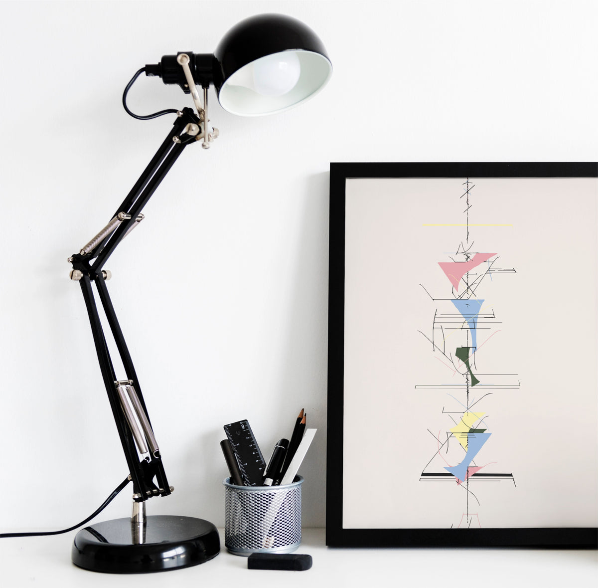 A black adjustable desk lamp with a white interior shade sits on a white surface next to a framed abstract art print. The lamp has a black base and silver metal joints, with a pen cup holding pens nearby.