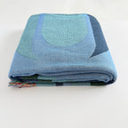 Folded blue and green woven blanket art piece with fringe on white background