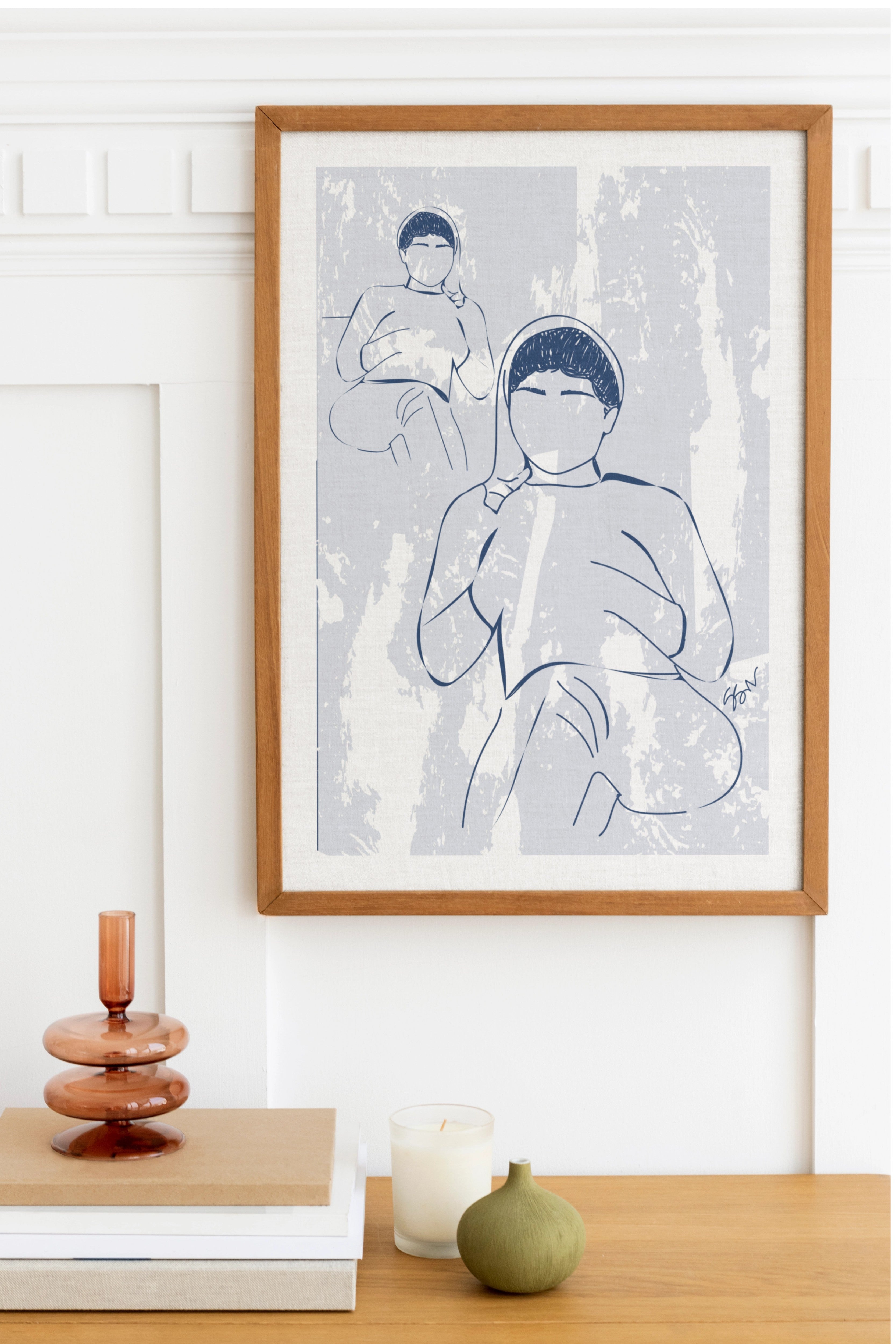 Framed line art print of two people on a wall above a wooden surface with decorative items.