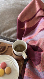 Red throw blanket covering person sitting on gray sofa with wooden tray with cup of tea and plate of cookies 