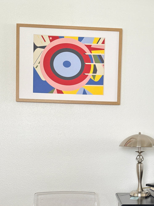 Colorful artwork in light brown frame on white wall.