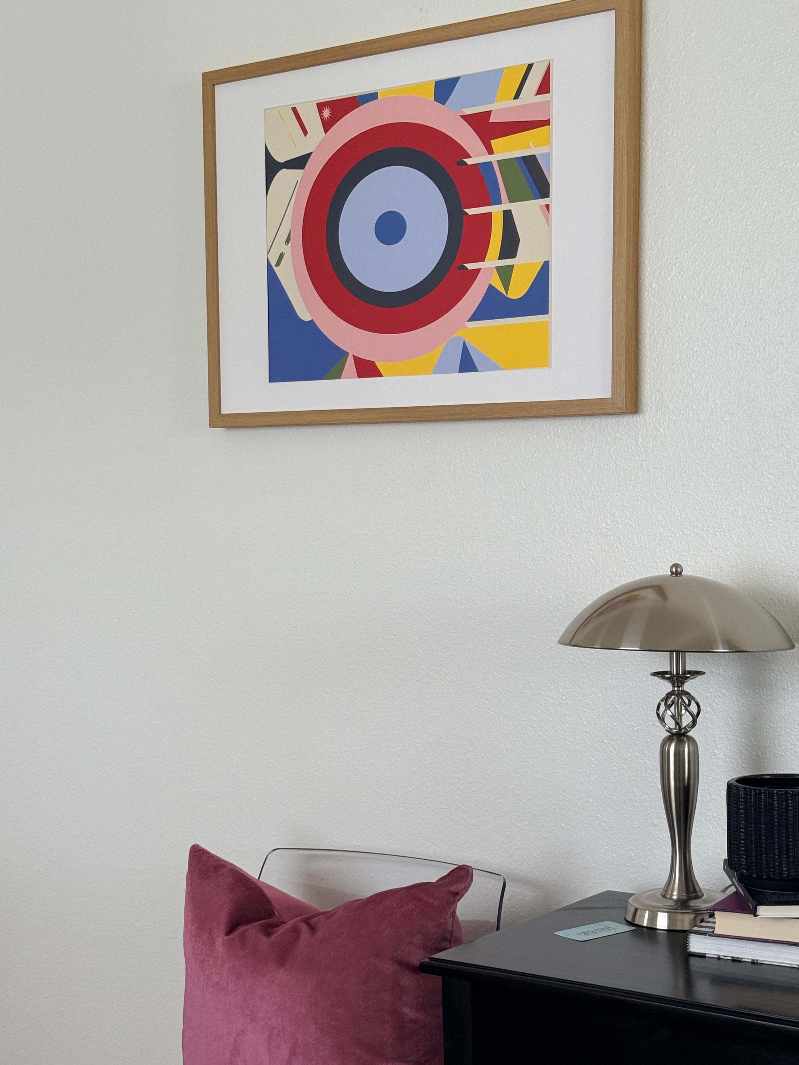 Framed abstract art on a white wall with a lamp and books on a shelf.