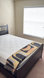 Abstract patterned black and yellow throw blanket draped over foot of queen sized dark bed with white sheets. 