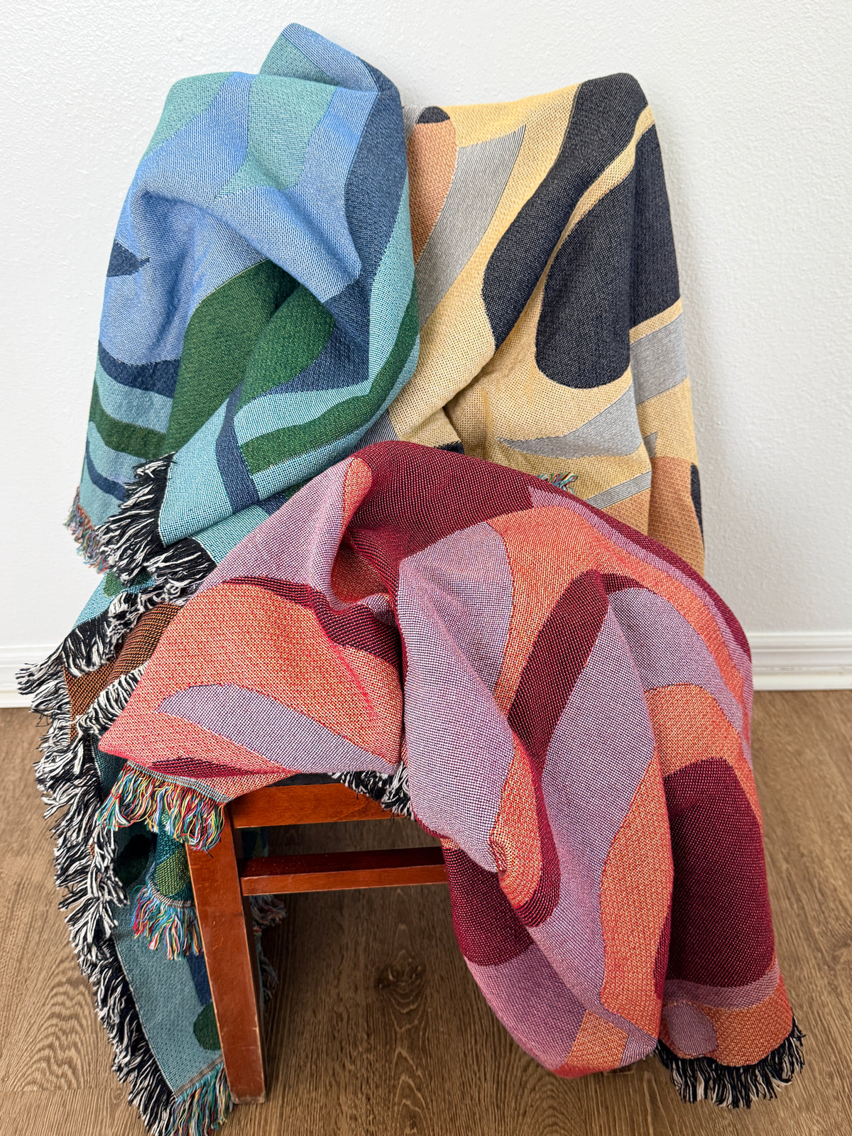 Three woven throw blankets with abstract geometric patterns in various colors are draped over a wooden chair in a room setting with a white wall and wood floor.