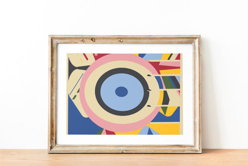 A wood framed abstract geometric art print with concentric eye-like shapes in blue, pink, yellow, and white, leaning against a beige wall on hardwood floor