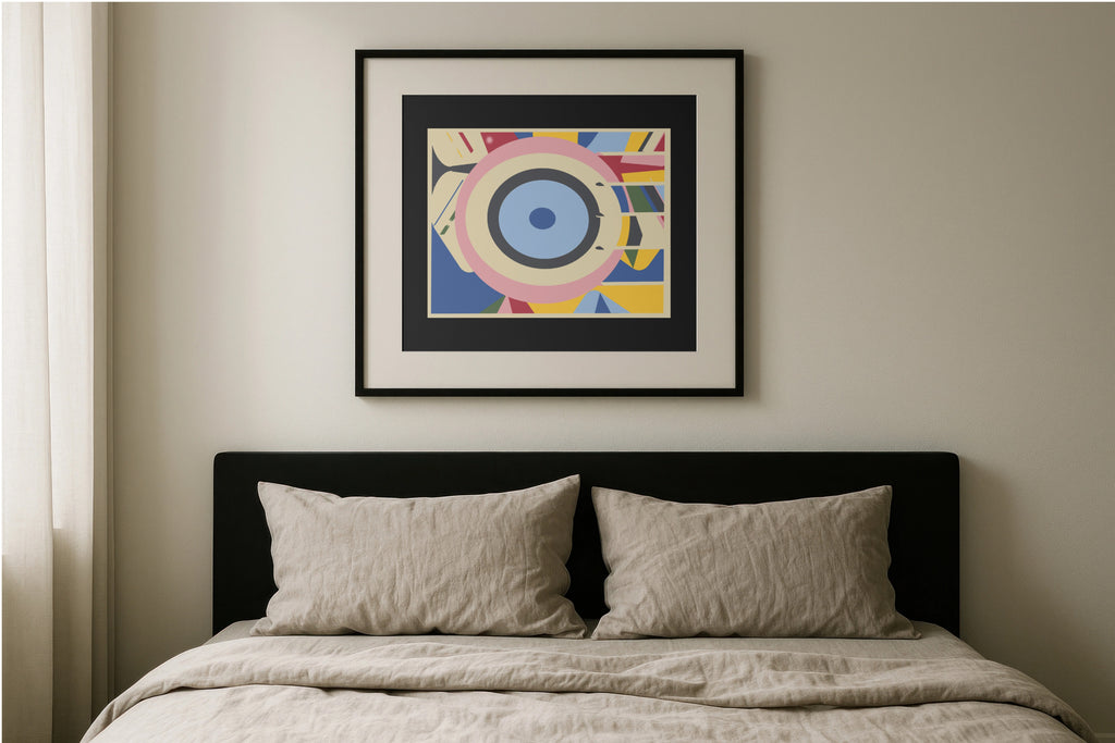A framed abstract geometric art print with concentric eye-like shapes in blue, pink, yellow, and white, hanging on a beige wall above a bed with a black headboard and beige bedding.
