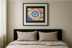 A framed abstract geometric art print with concentric eye-like shapes in blue, pink, yellow, and white, hanging on a beige wall above a bed with a black headboard and beige bedding.
