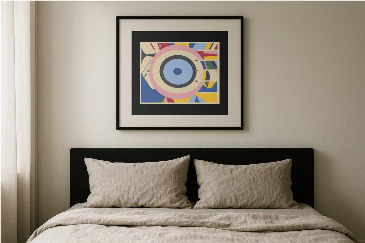 A framed abstract geometric art print with concentric eye-like shapes in blue, pink, yellow, and white, hanging on a beige wall above a bed with a black headboard and beige bedding.