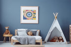 A tan framed abstract geometric art print with concentric eye-like shapes in blue, pink, yellow, and white, hanging on a blue wall in kid room with wood bed and white tent with stuffed animals.